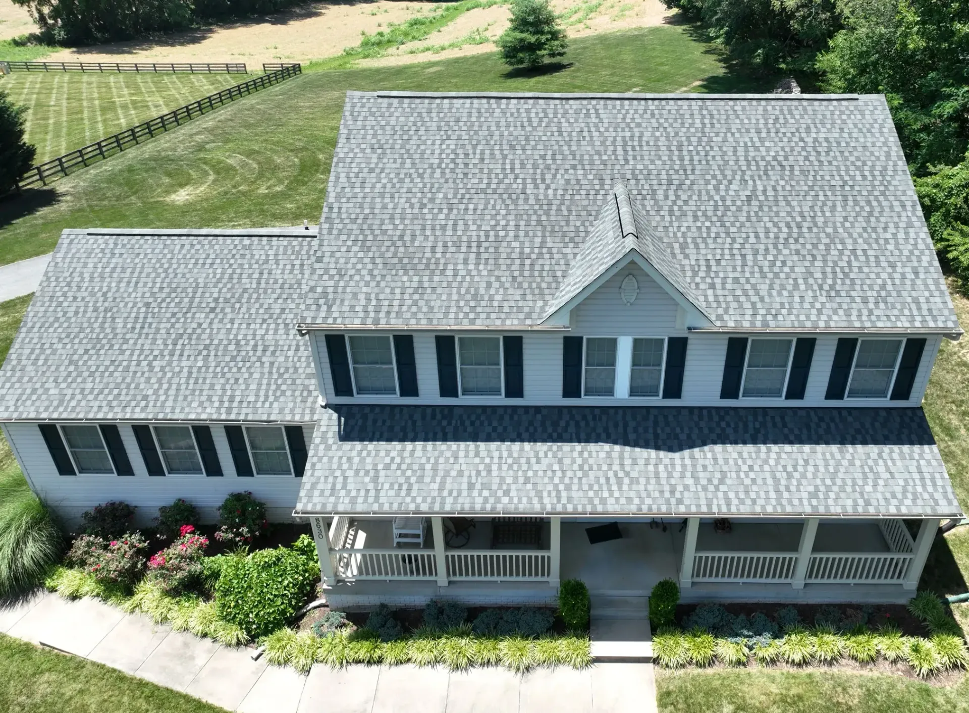 Aerial view of a two-story house with a gray shingle roof, light-colored siding, black shutters, and a covered porch, set on a large grassy lot