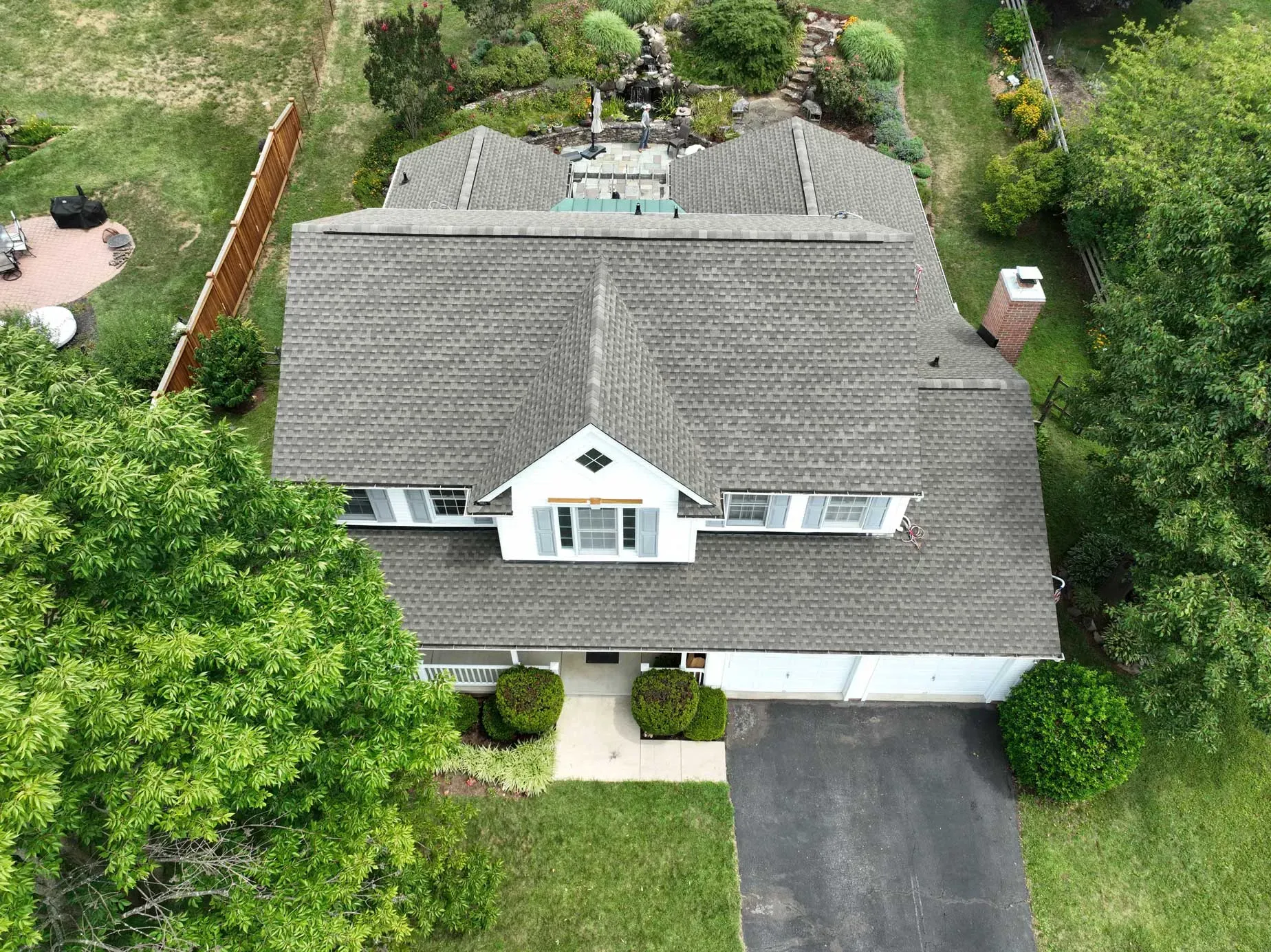 Aerial view of a large white farmhouse with a new gray shingle roof, with a large amount of shrubs and trees circling the property