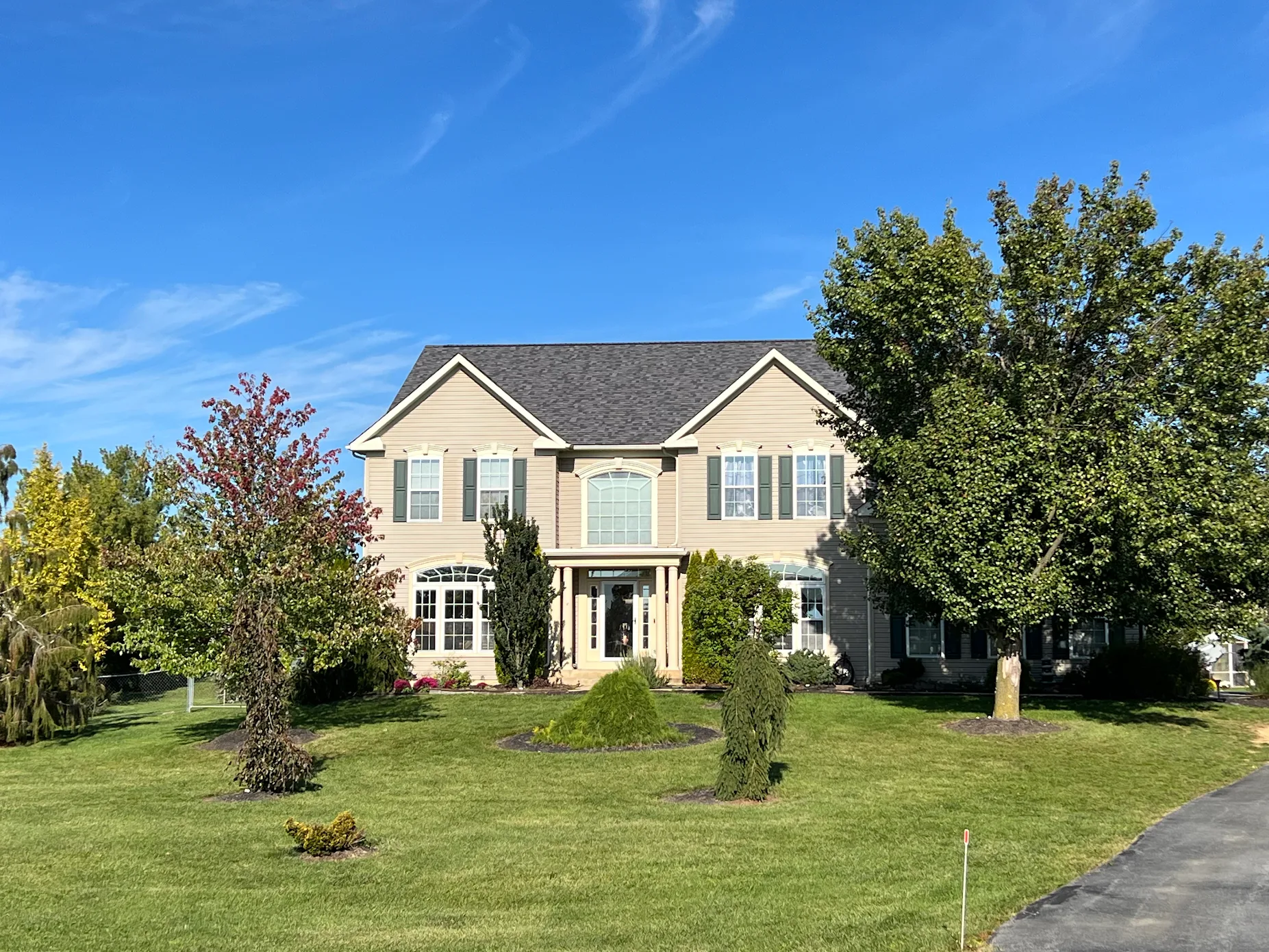 Two-story beige house with a dark gray roof, green shutters, and a large arched window above the front door, surrounded by a green lawn, mature trees, and a clear blue sky