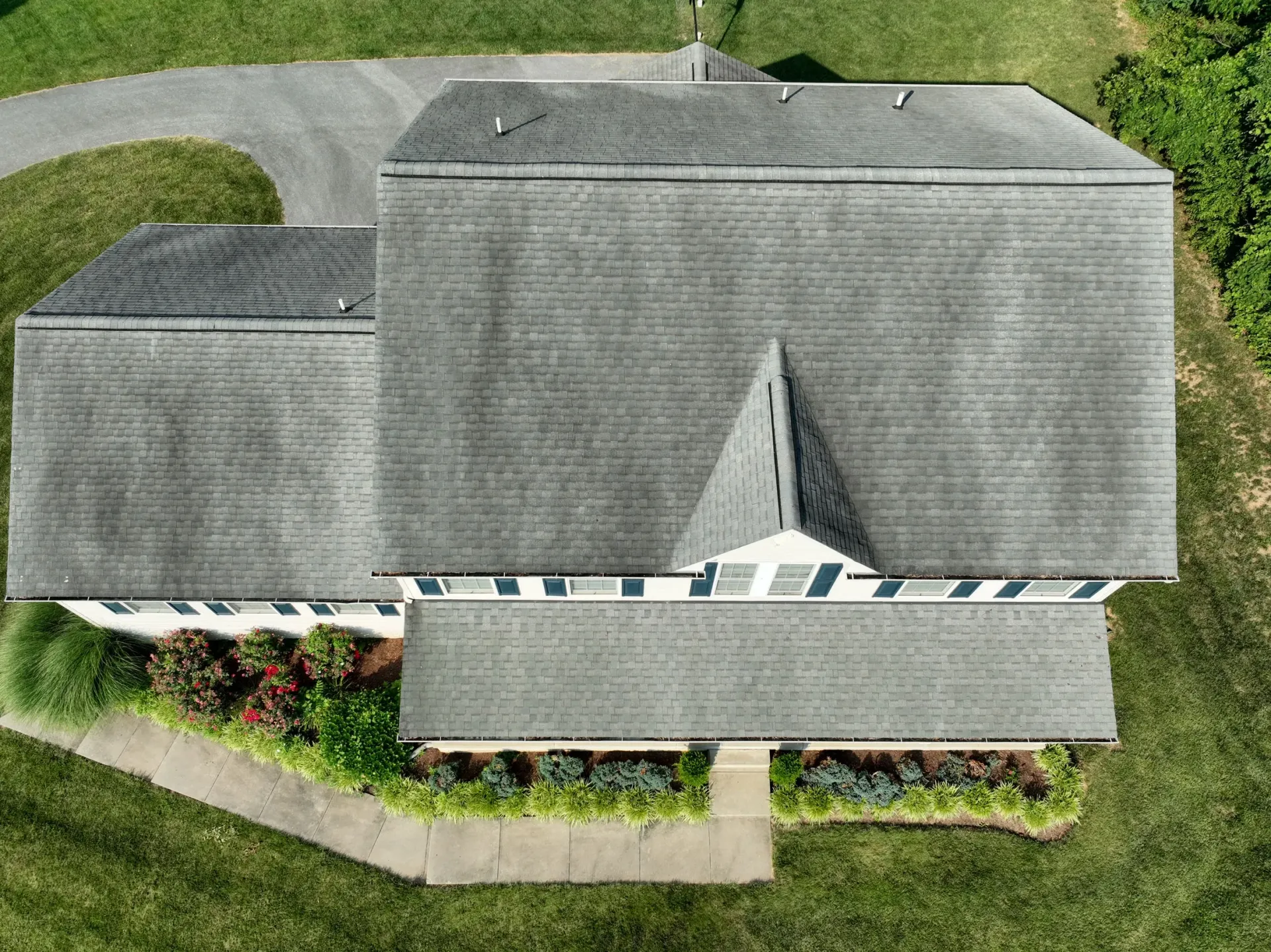 Aerial view of a worn gray roof on a 2-story white farmhouse surrounded by green grass and colorful landscaping