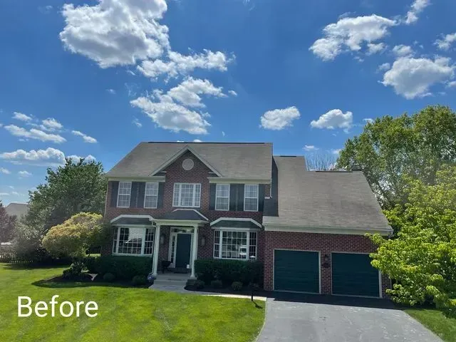 Two-story red brick house with green shutters and garage doors, set against a bright blue sky with scattered white clouds, surrounded by green foliage
