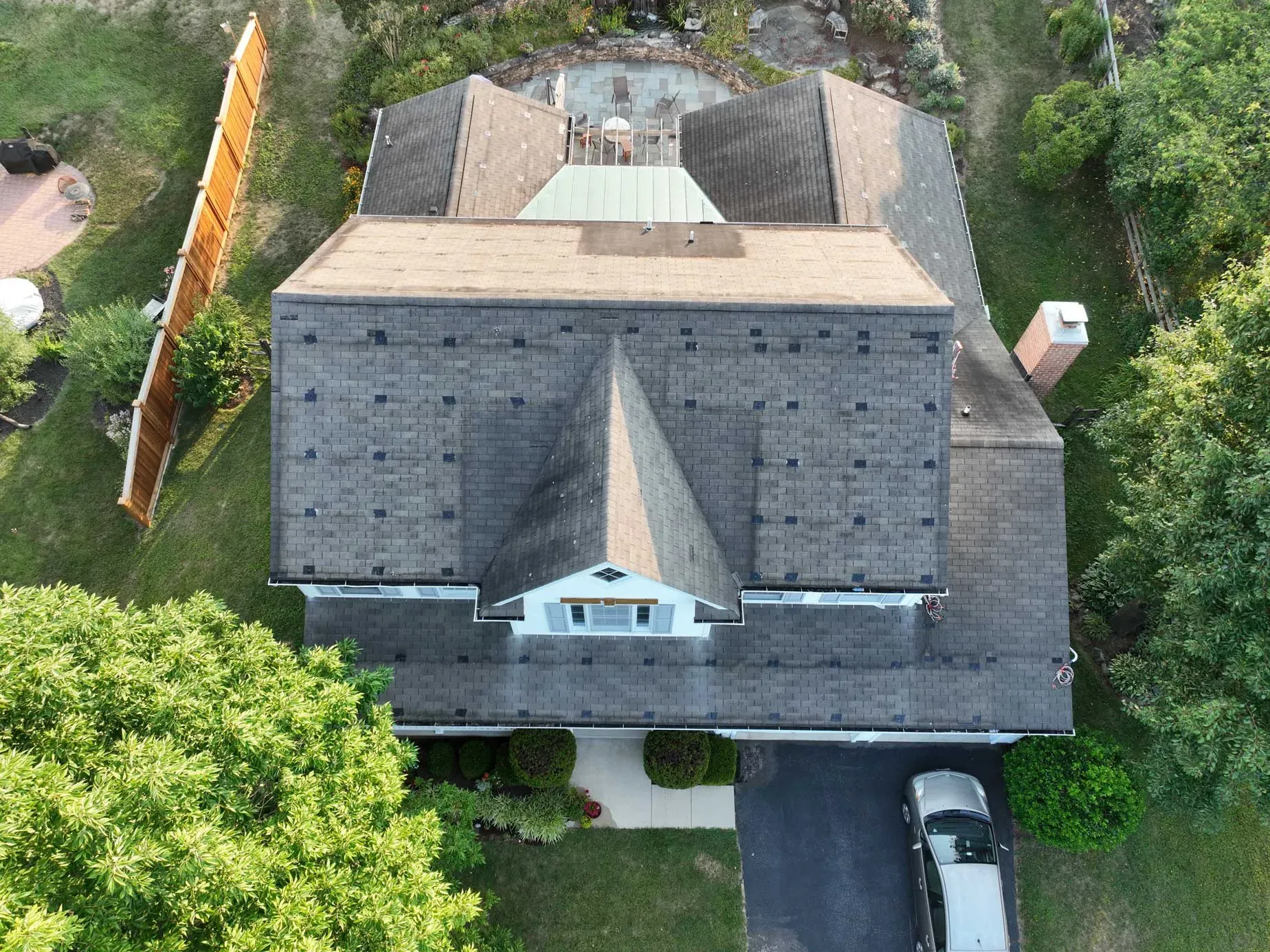 Aerial view of a multi-sectioned house with a complex roof structure composed of grey shingles, a white dormer window with shutters, a brick chimney, and surrounding greenery including trees, a lawn, and a wooden fence; a car is parked in the driveway
