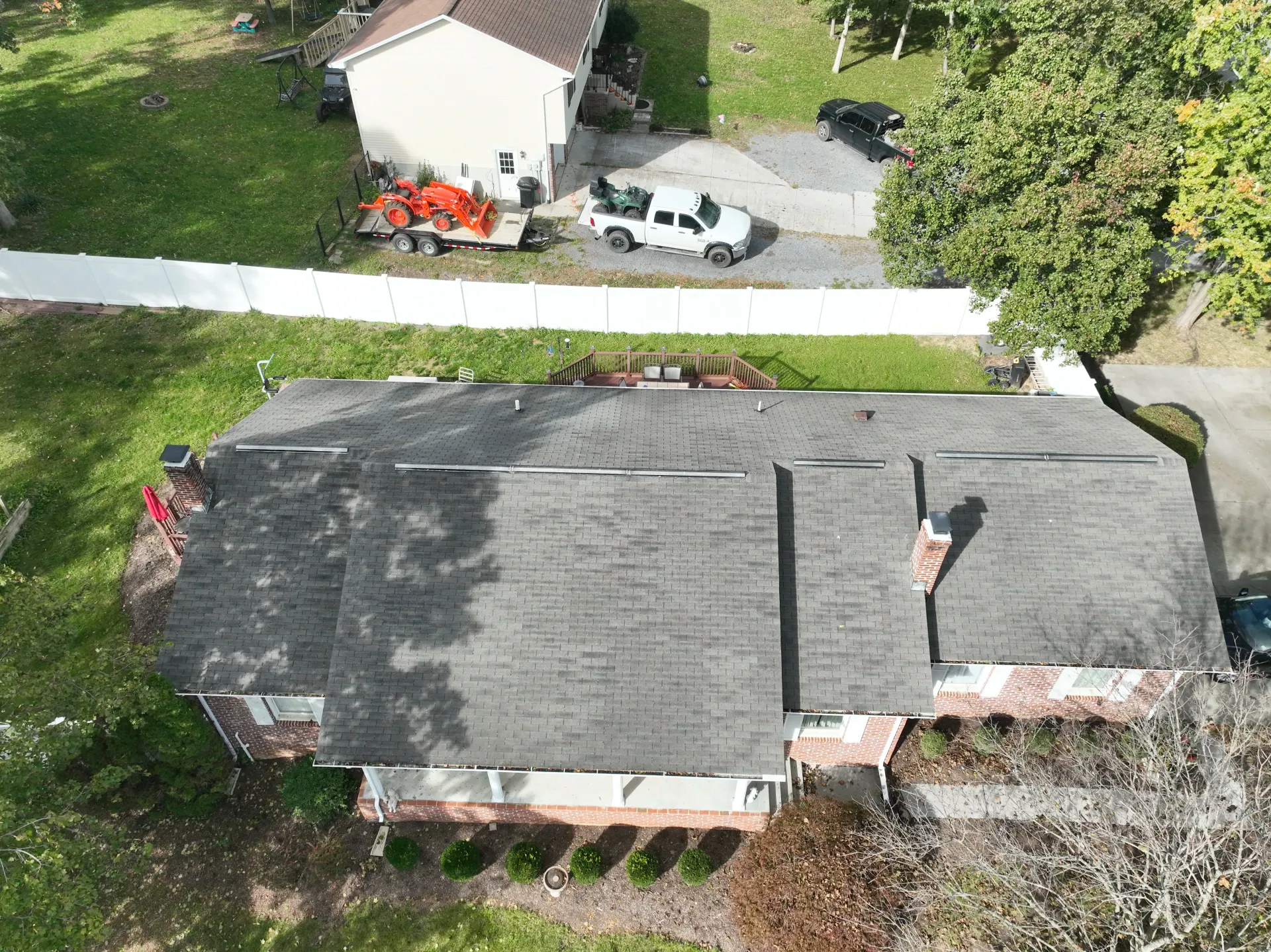 Aerial view of a suburban house with a worn gray shingle roof, chimney, and adjacent white fence, driveway, and surrounding properties, including a white pickup truck, a tractor on a trailer, and a black vehicle