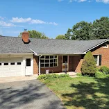 A red brick single story home with a red brick chimney, gray shingles, white garage door and a large patch of grass in the front