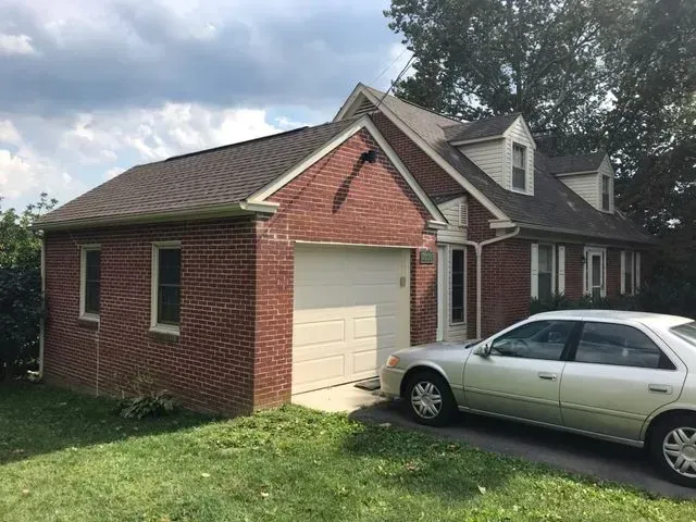 Red brick house with a detached garage and a silver sedan parked in the driveway. The house has a brown shingle roof, white trim, and two dormer windows. The garage has a white garage door. Green grass and trees surround the house.
