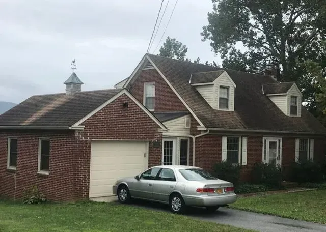 A two-story red brick house with a worn roof, detached garage and dormers, with a silver sedan parked in the driveway, and topped with a weather vane