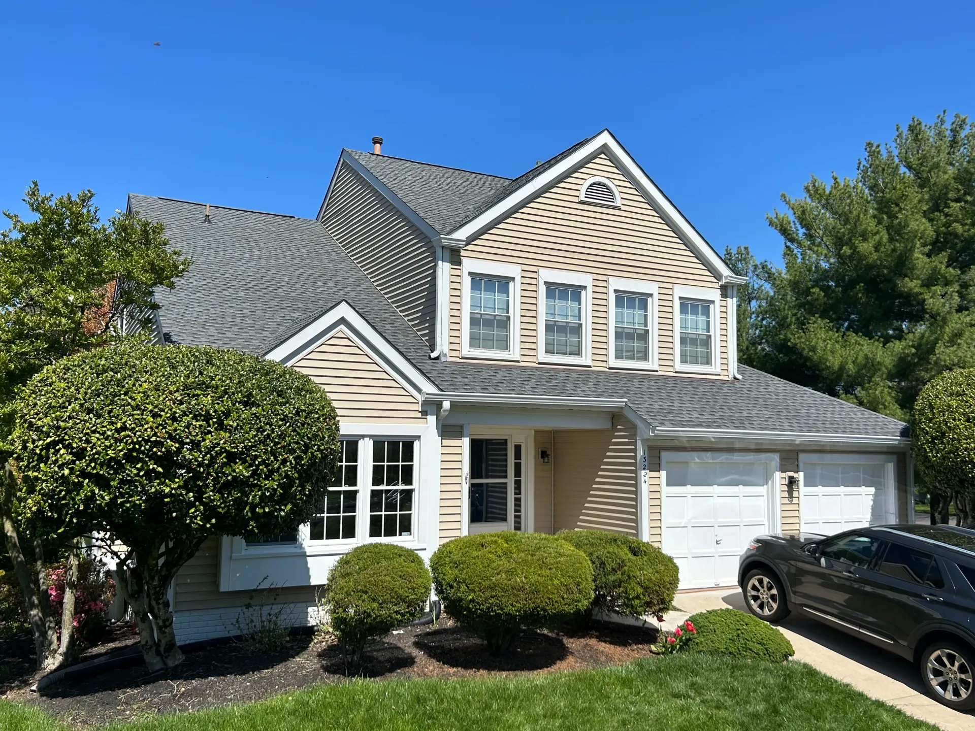 Two-story beige house with gray shingle roof, white trim, three upper windows, and two garage doors, surrounded by green shrubs, trees, and a dark SUV parked in the driveway under a clear blue sky