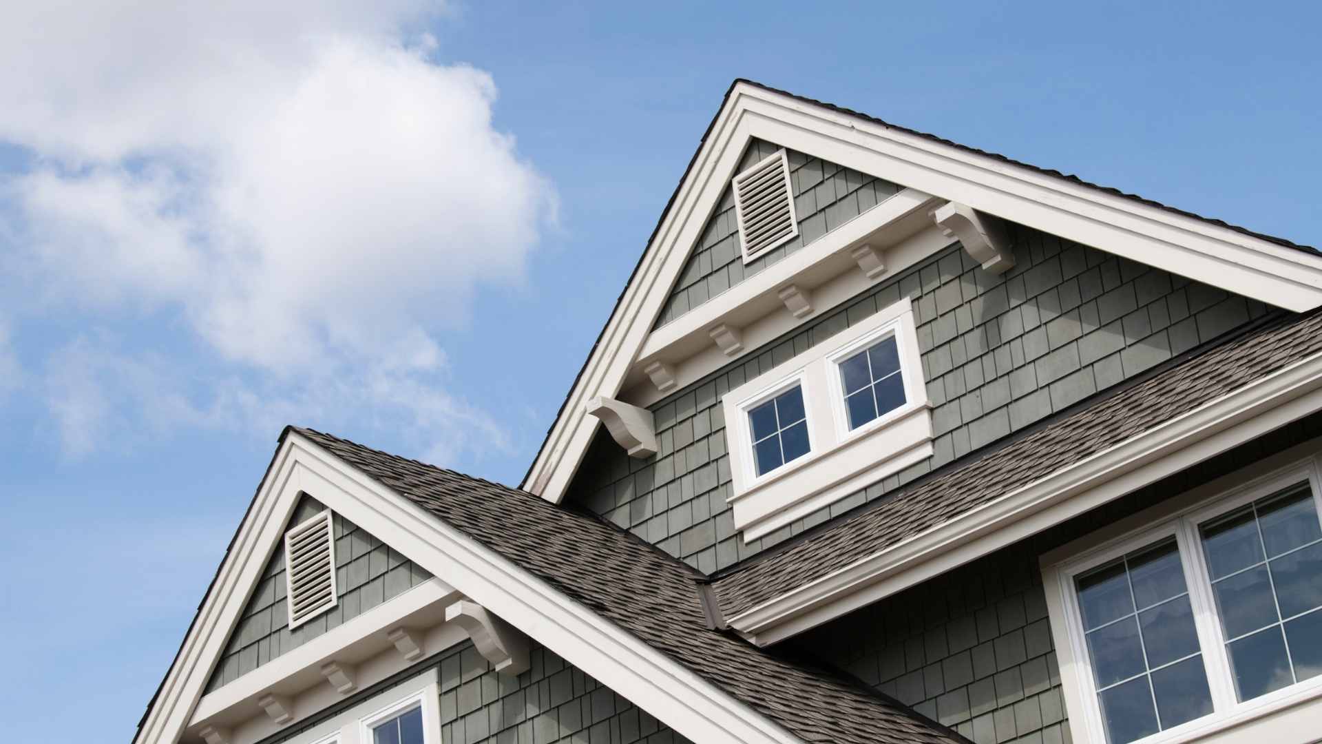 Close-up of a grey shingled house roof with white trim, gables, and dormer windows against a blue sky
