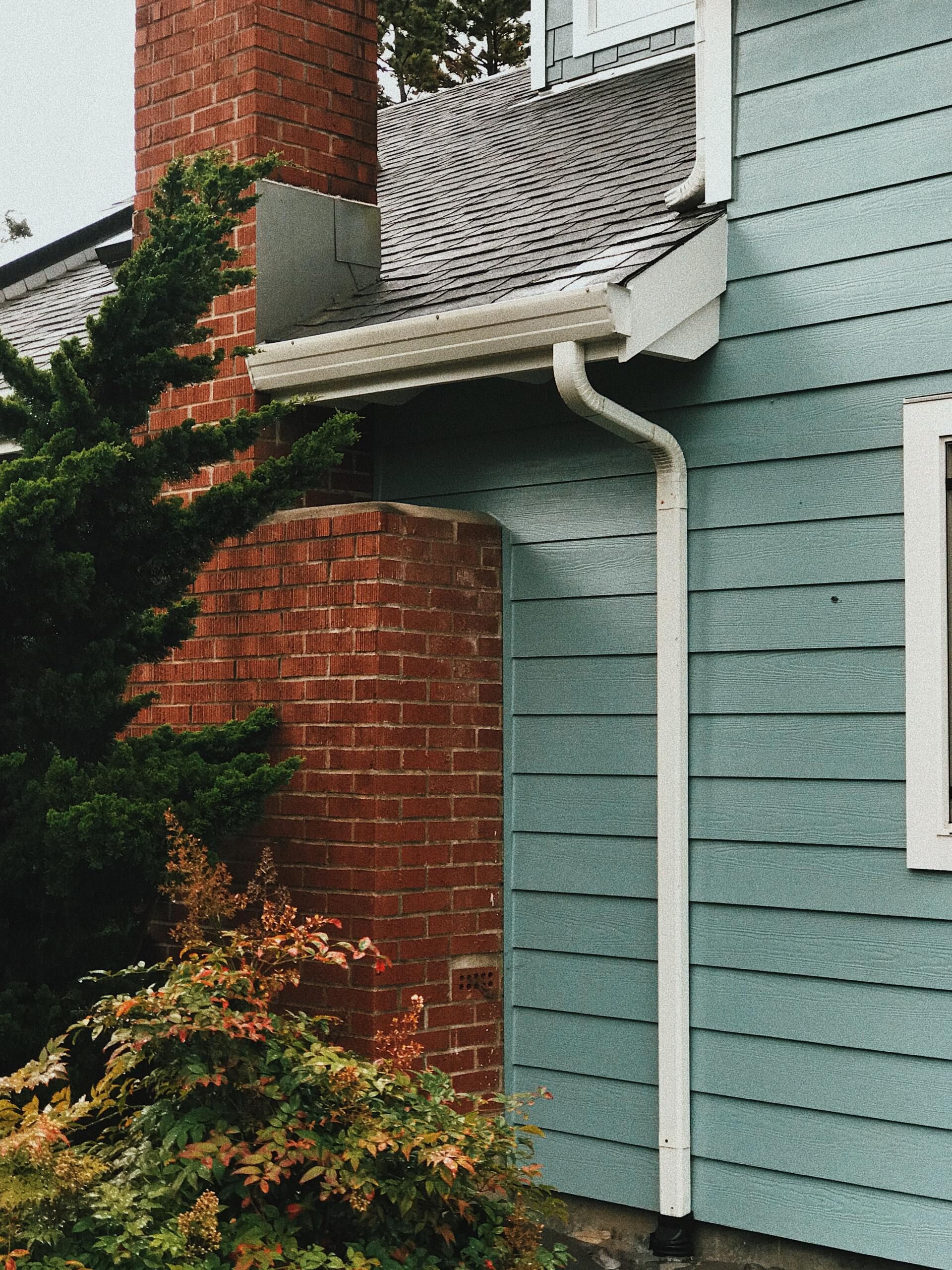 Exterior of a house with a red brick chimney and teal siding - a white gutter runs along the roofline, connected to a downspout on the side of the house and the house is surrounded by fall trees and foliage