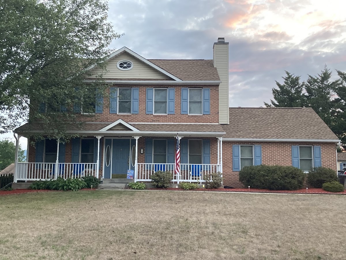 Two-story brick house with blue shutters and a front porch, an American flag hanging on the porch, a large tree to the left, and a cloudy sunset sky