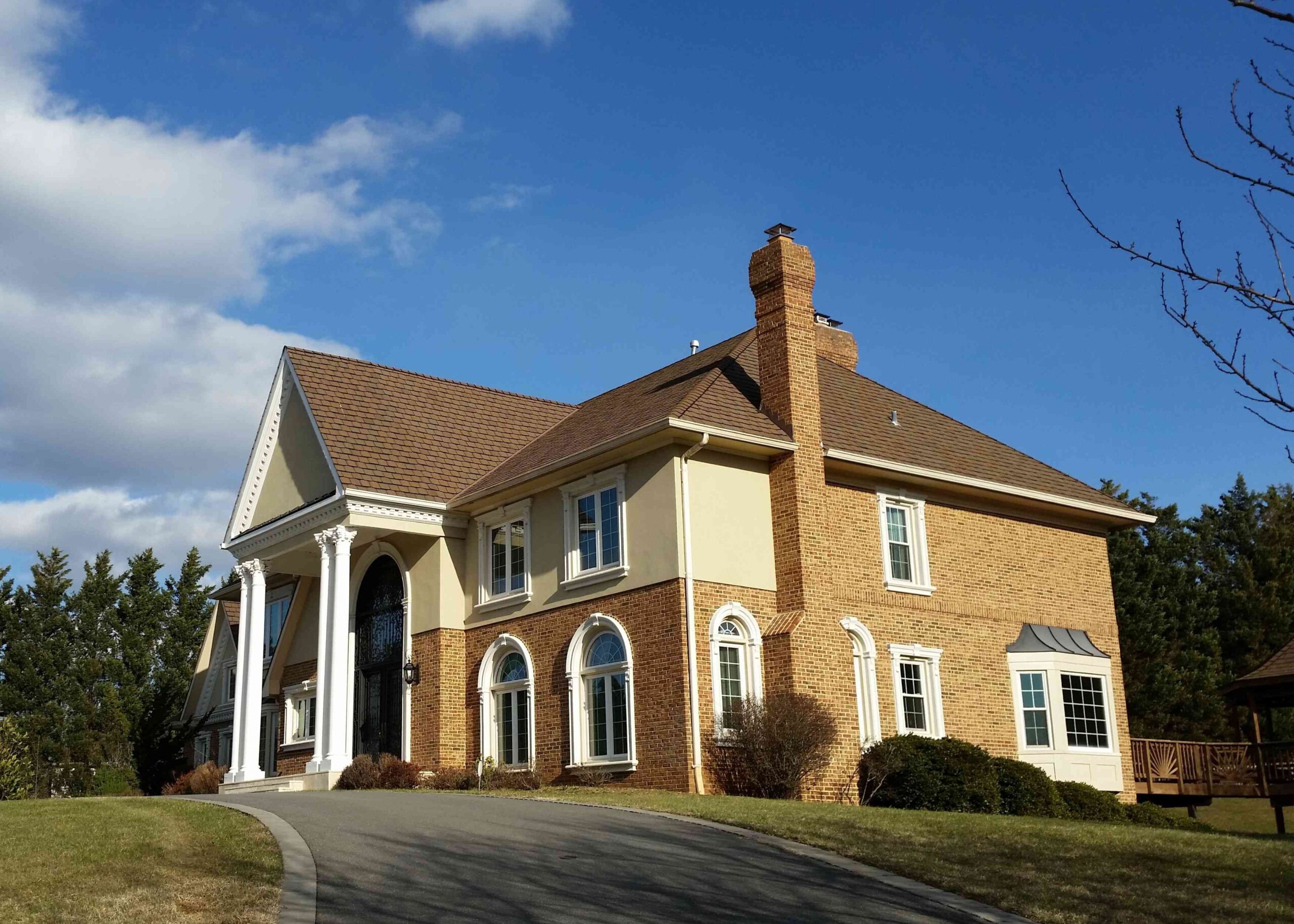 Large brick home with a tall chimney, white columns near the front door, and mixed brown-red shingle roofing