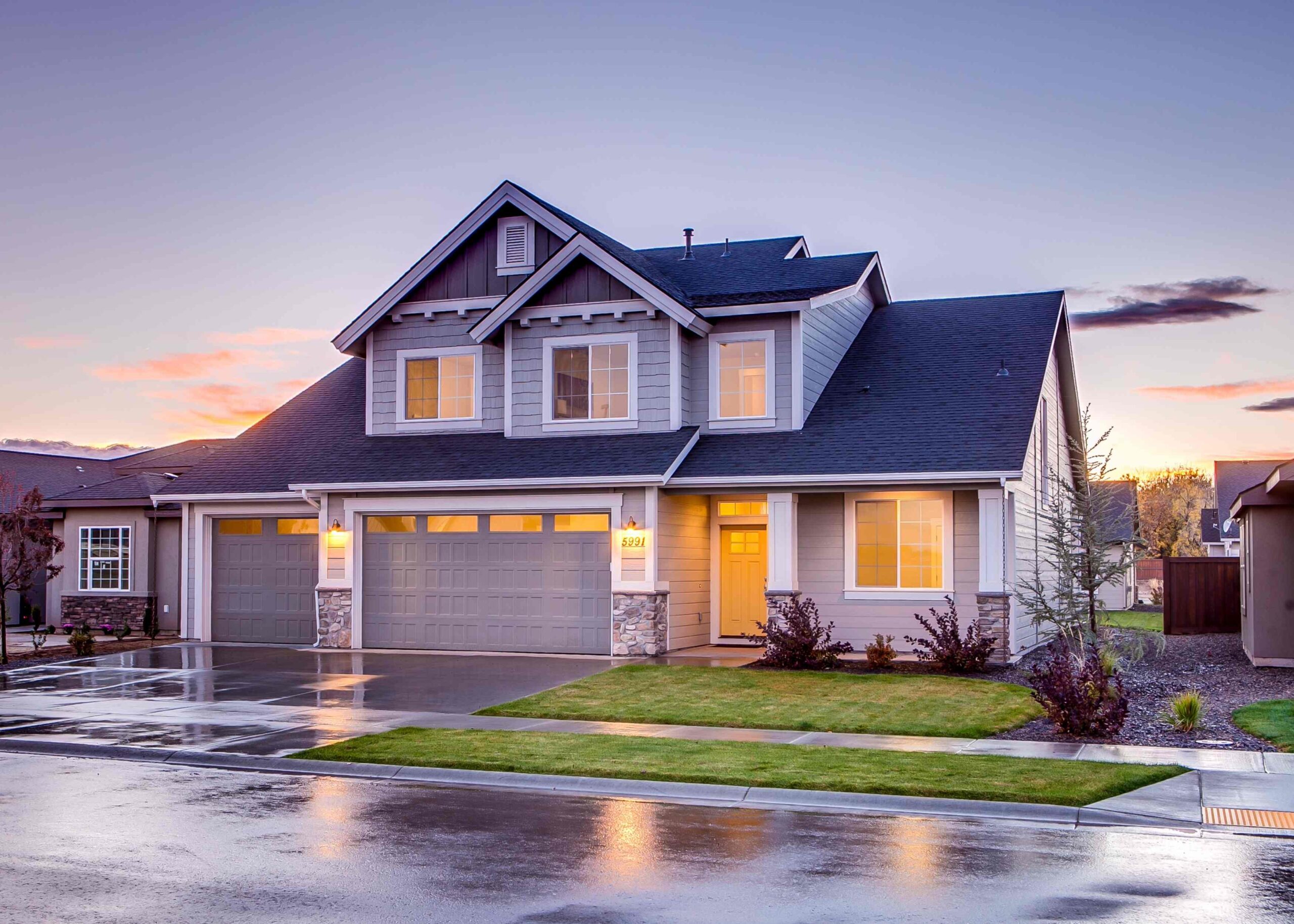 Modern 2-story home with a dark blue roof, lit from the inside - the driveway and street are wet from rain