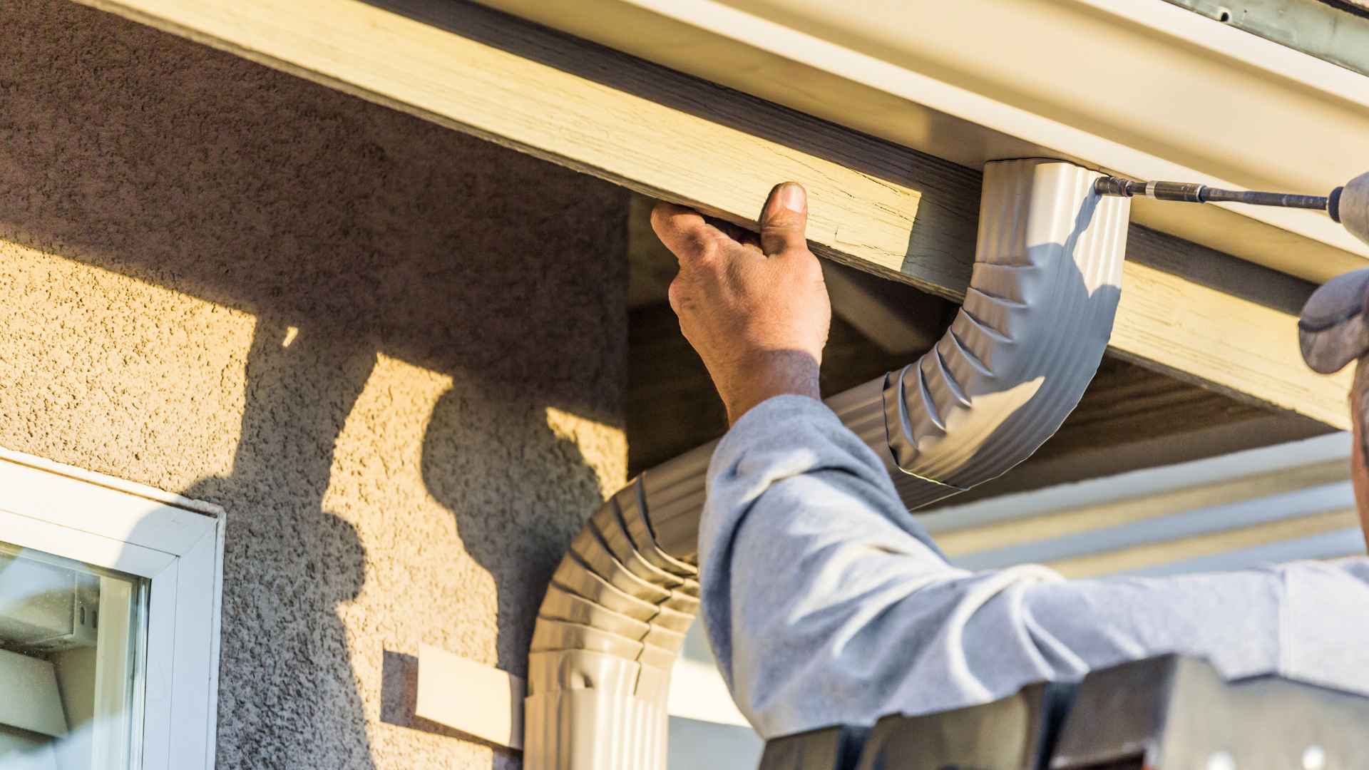 A man holding up a rain gutter with his left hand and screwing the gutter into the house with his right hand