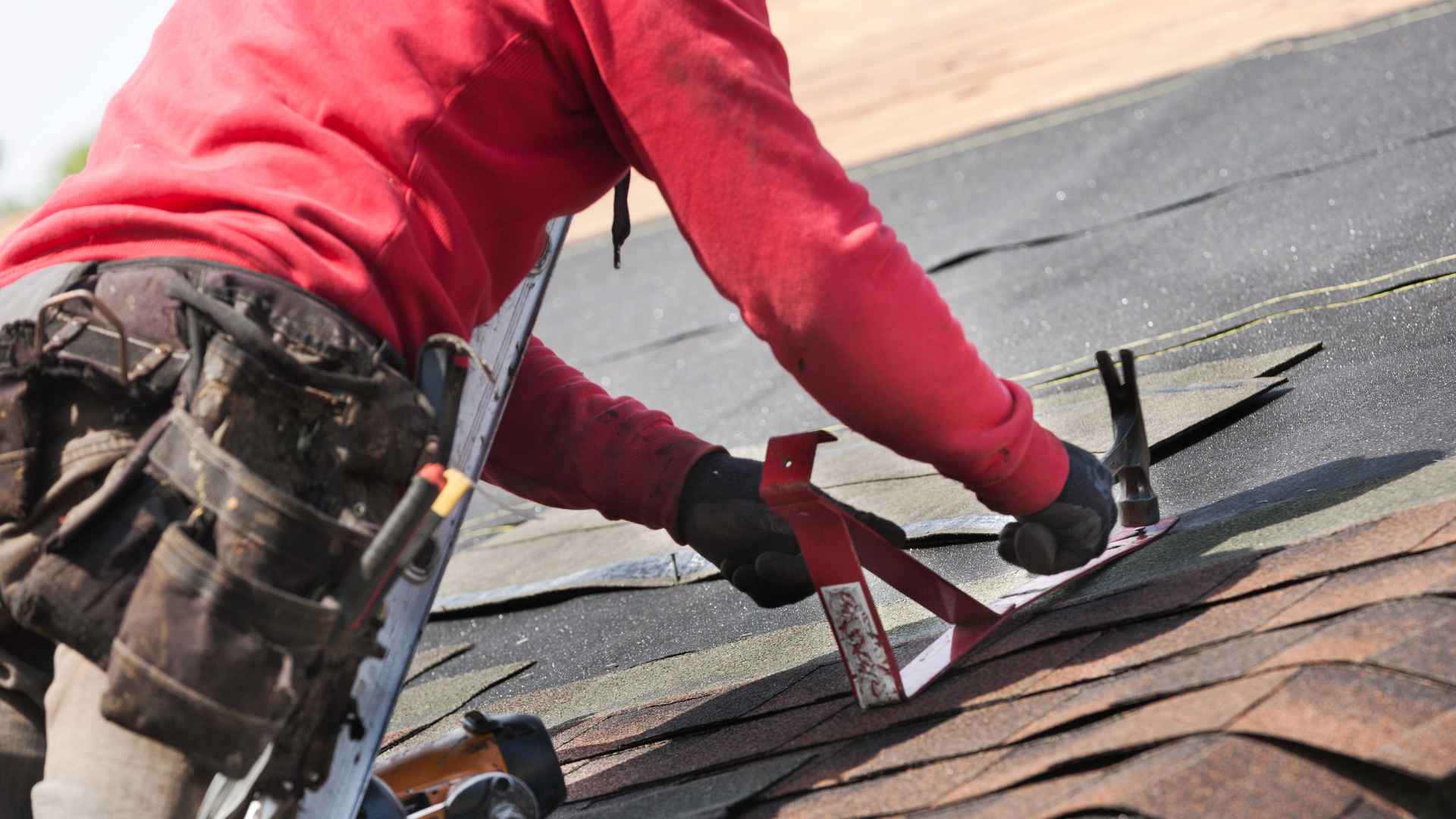 A man wearing a red shirt, engaged in roofing work and hammering shingles down