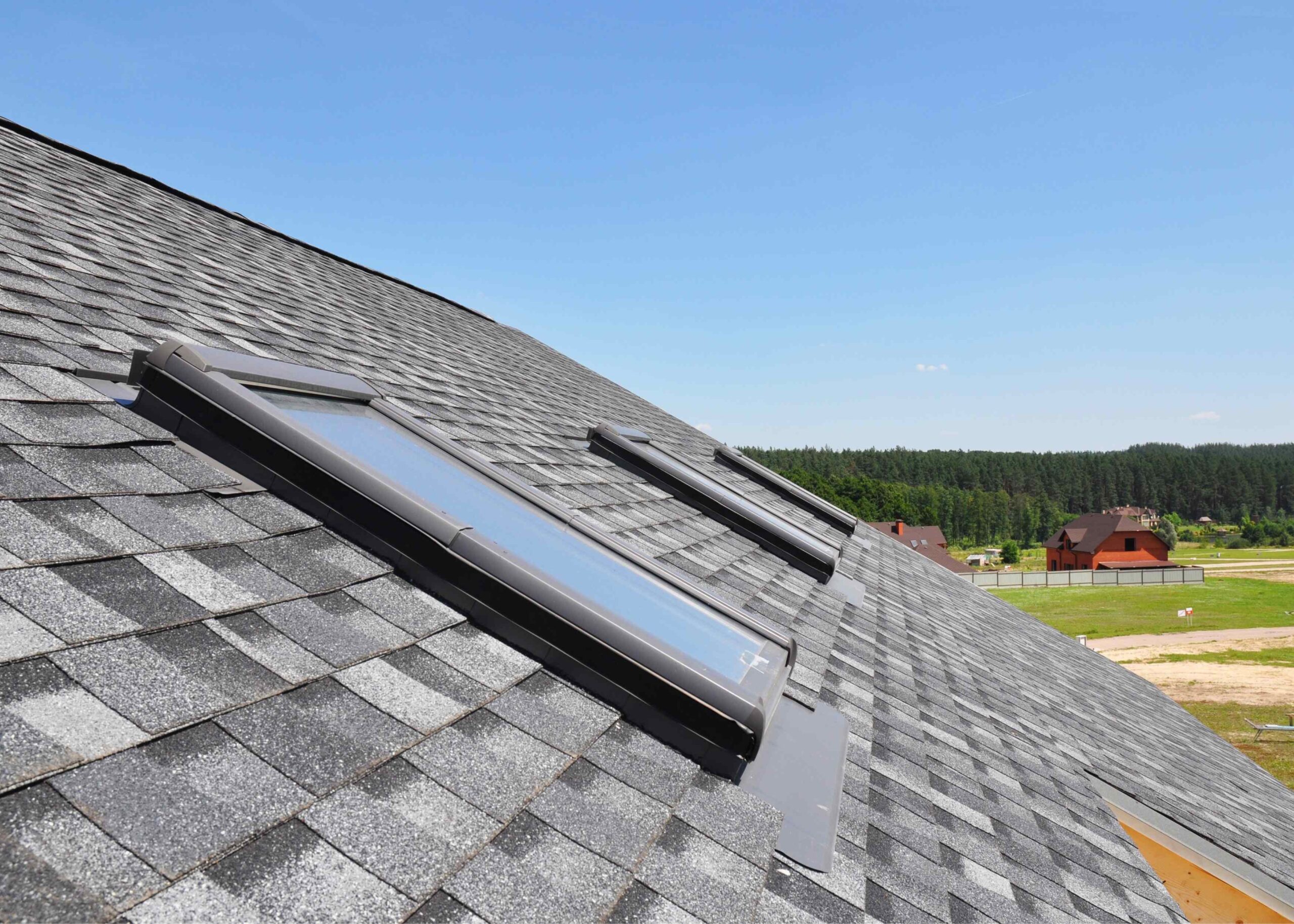 Close-up of a grey asphalt shingled roof with multiple skylights installed, overlooking a rural landscape
